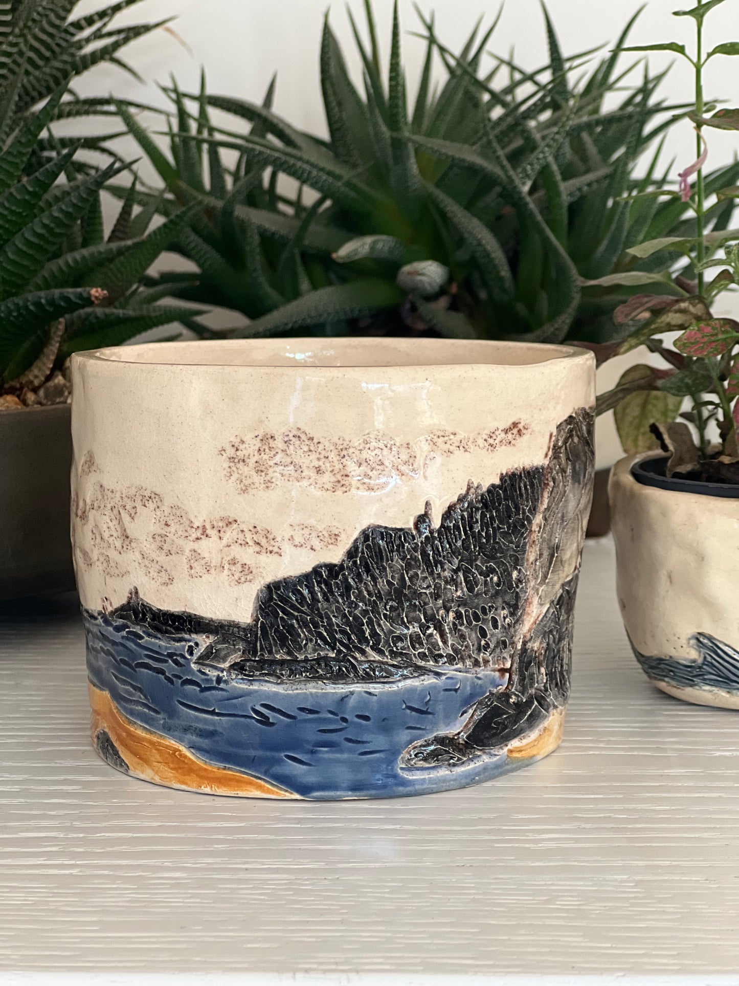 A handcrafted stoneware pot with a coastal landscape design, featuring elements of land and sea, placed on a table beside a potted plant.