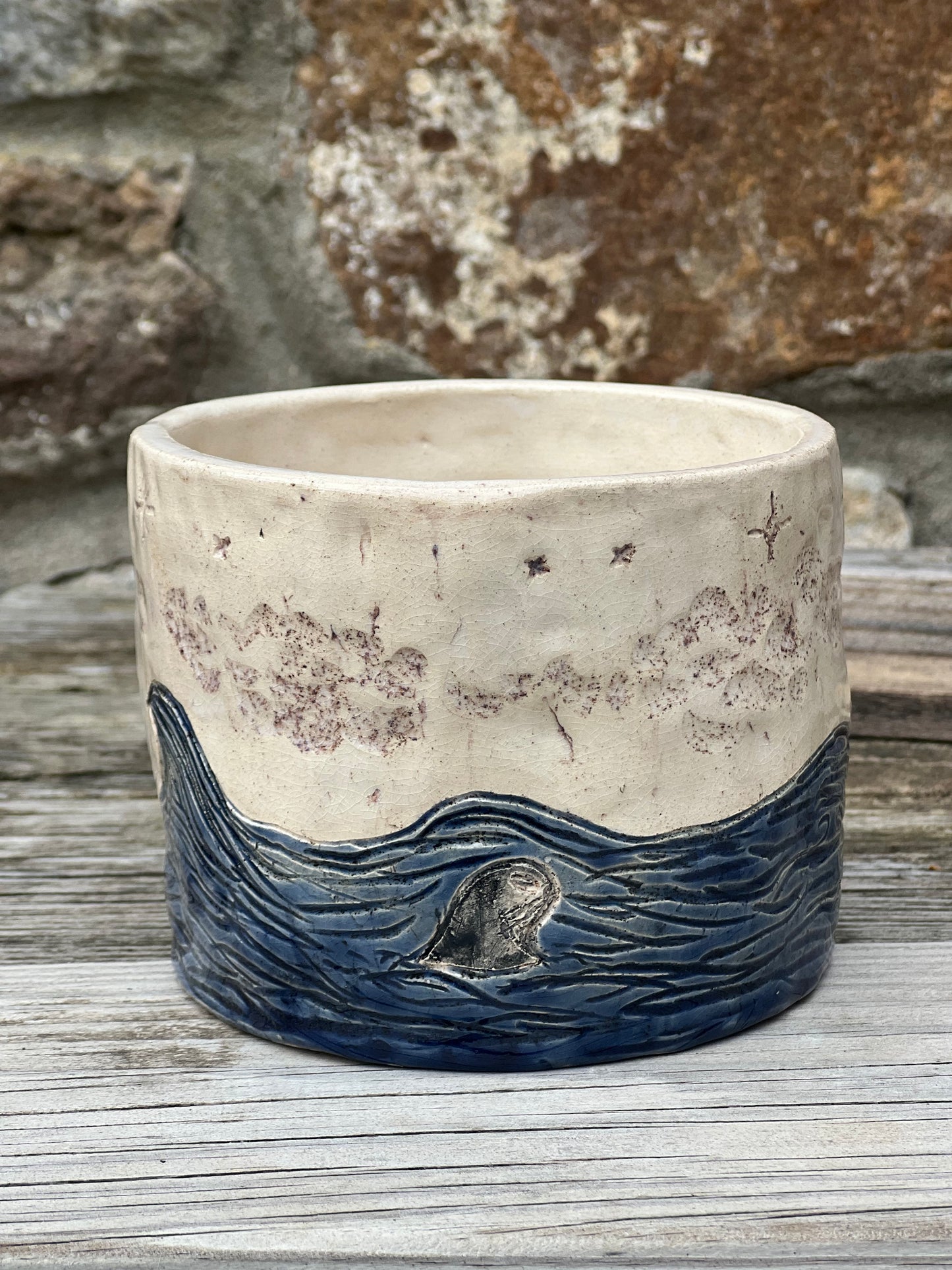 Handcrafted ceramic pot with a design of blue waves and a seal, under a star-speckled sky, on a wooden surface.