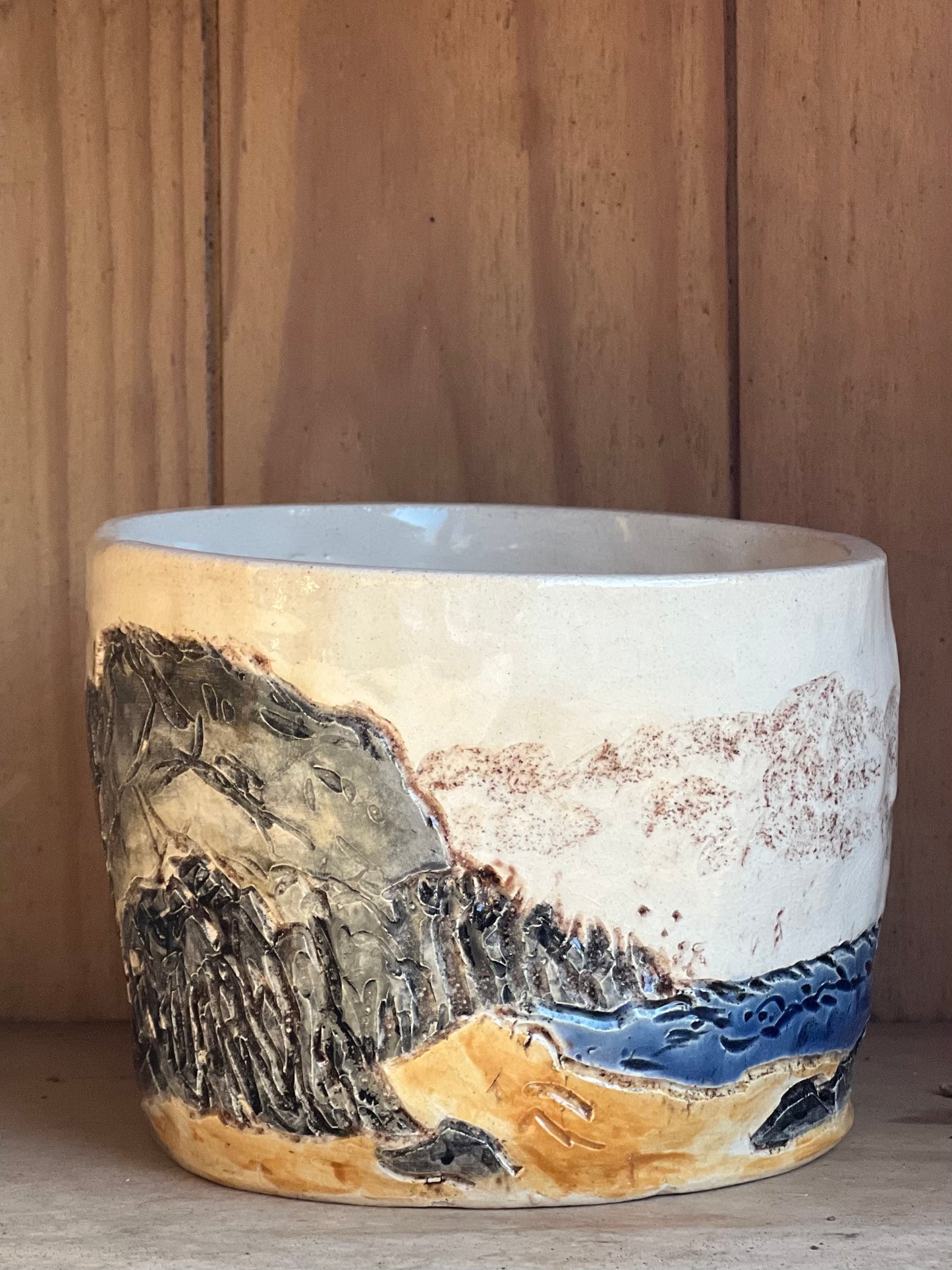 A handcrafted stoneware pot with a rustic design, featuring carved textures and a layered glaze that resembles a coastal landscape with cliffs and waves.