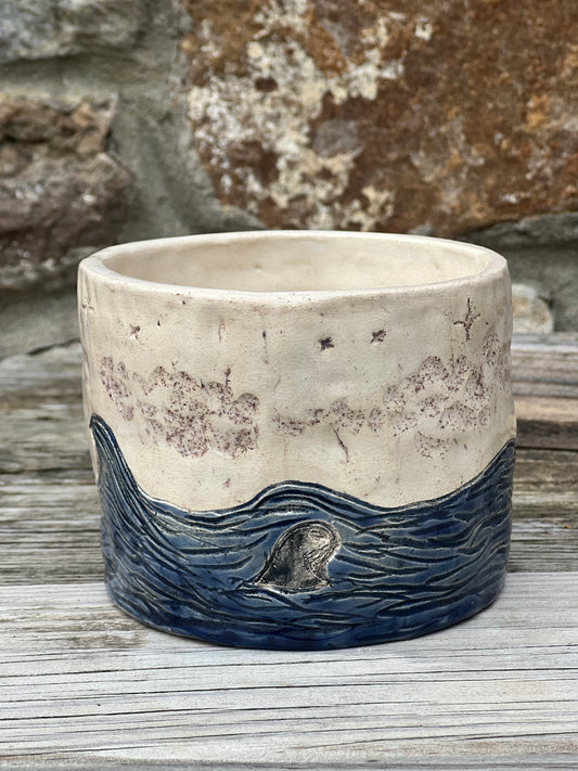 Handcrafted ceramic pot with a design of blue waves and a seal, under a star-speckled sky, on a wooden surface.